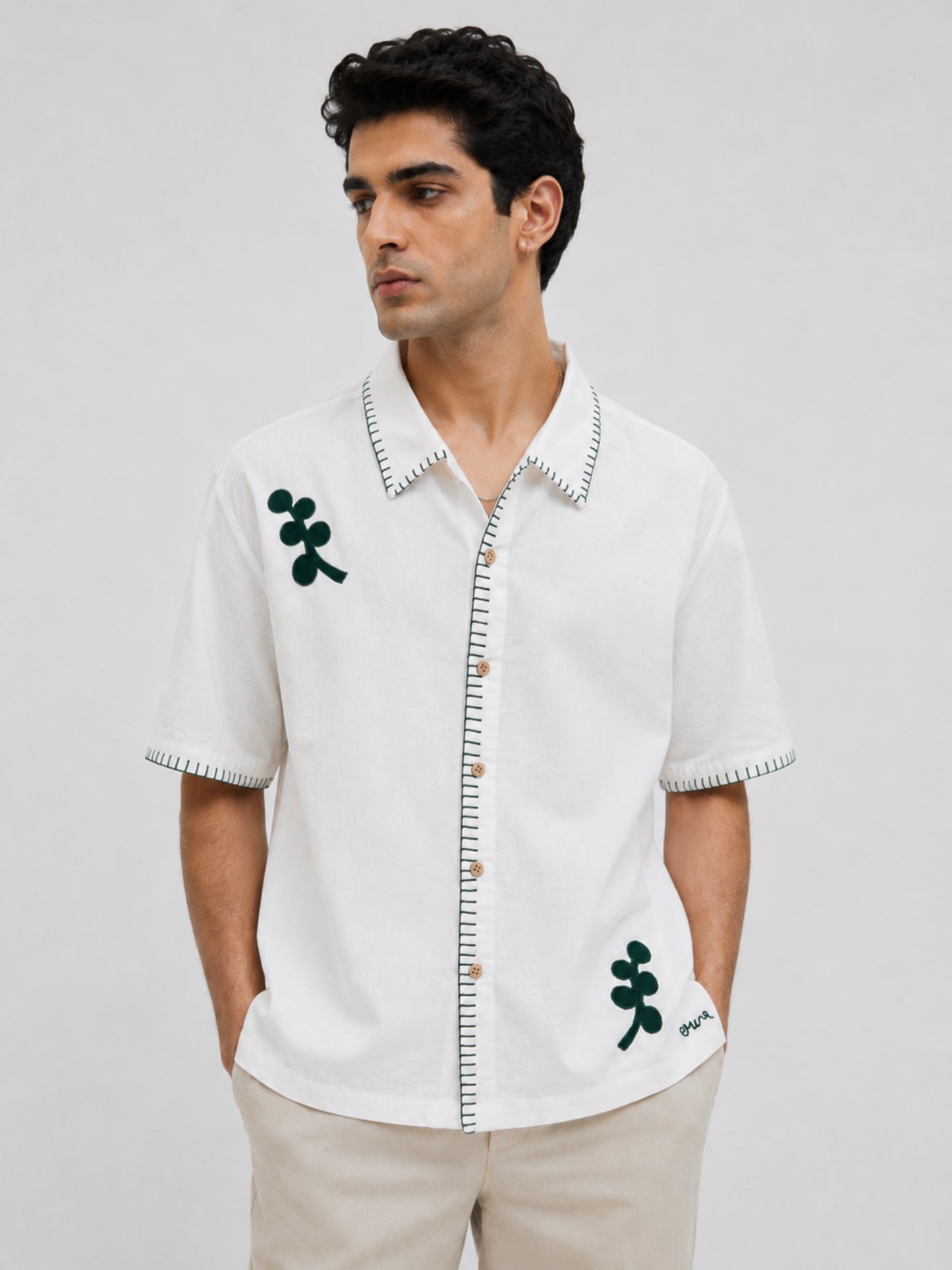 A man wearing a white short-sleeve button-up shirt with green embroidered motifs and black-and-white striped trim, paired with beige trousers, against a light gray studio background.