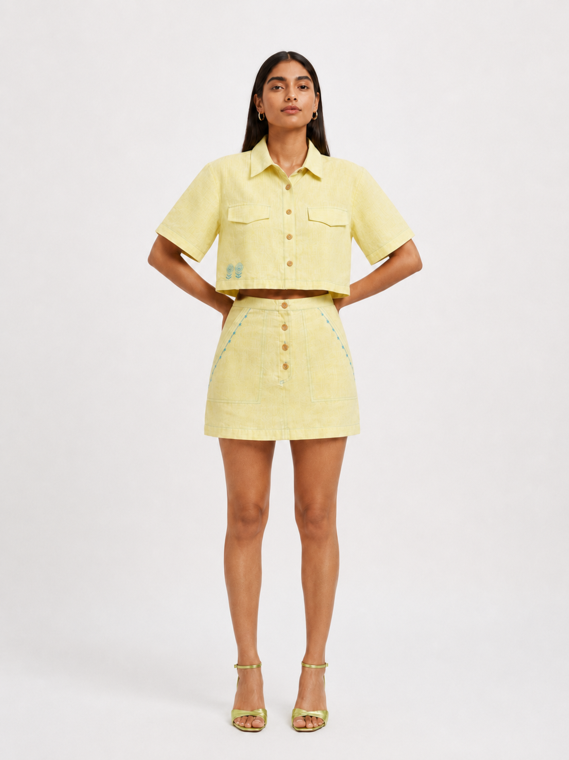 A model stands against a light gray studio background wearing a lemon yellow short-sleeve button-front shirt and matching mini skirt with small embroidered details, paired with metallic gold high-heeled sandals.