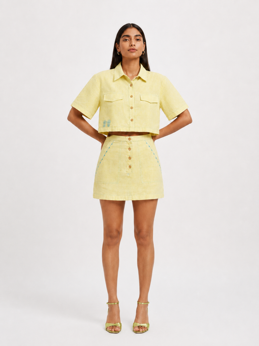 A model stands against a light gray studio background wearing a lemon yellow short-sleeve button-front shirt and matching mini skirt with small embroidered details, paired with metallic gold high-heeled sandals.