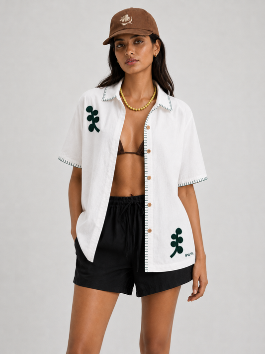 A model wearing a white short-sleeve button-up shirt with green embroidered leaf motifs and black shorts, paired with a brown baseball cap and gold necklace, against a light gray studio background.